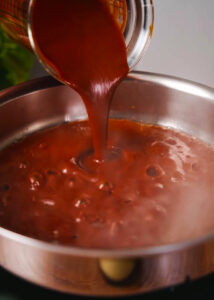 Salsa roja being poured into a stainless steel pan with hot rendered pork fat