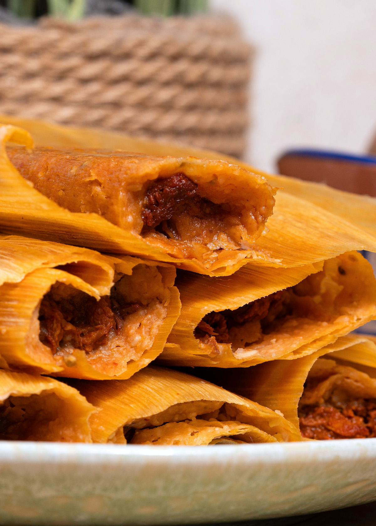 A plate of Red Pork Tamales with their corn husks partially open reveals a spicy meat filling. The tamales are stacked, showcasing the savory red filling inside the golden corn dough.