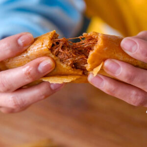 A close-up of hands pulling apart a tamale, revealing its savory filling. The tamale is wrapped in a golden corn husk, and the filling looks like shredded meat. The background is blurred, showing a person in a blue and yellow shirt.