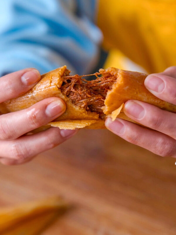 A close-up of hands pulling apart a tamale, revealing its savory filling. The tamale is wrapped in a golden corn husk, and the filling looks like shredded meat. The background is blurred, showing a person in a blue and yellow shirt.