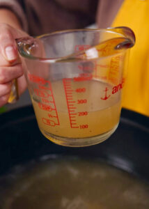 A person holds a glass measuring cup filled with pork broth, marked with red measurement lines up to 400 milliliters.