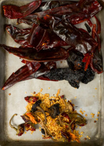 An overhead view of various dried chili peppers and seeds on a metal baking tray. The peppers are deseeded and stems removed.