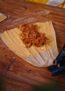 Masa (corn dough) spread on a corn husk with red pork fillings.