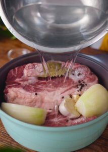 A pot filled with a raw pork butt, cut onions, garlic cloves, and black peppercorns. A saucepan is pouring water over the ingredients, preparing for cooking.