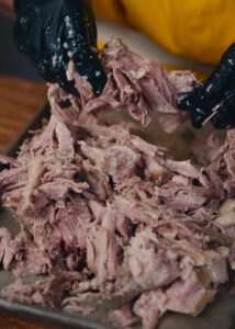 A person wearing black gloves pulls apart tender, cooked shredded meat on a tray. The meat appears juicy and succulent.