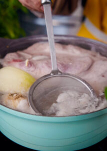 A ladle skims foam off a simmering broth in a pot, revealing bone-in pork butt, an onion, and garlic cloves beneath the surface. The pot is part of a cooking process.