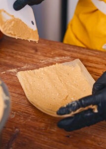 A person wearing black gloves spreads masa dough onto a corn husk, in preparation for making tamales.