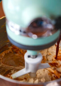 Close-up of a light blue stand mixer blending dough in a stainless steel bowl. The mixing paddle is in motion, creating a blurred effect. The dough appears partially mixed with streaks of a darker substance, possibly chocolate or sauce.