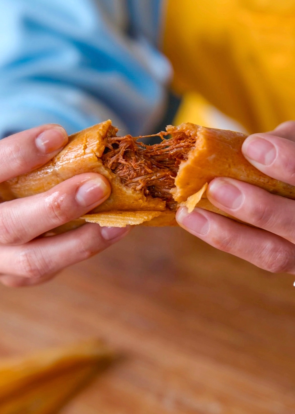 A close-up of hands pulling apart a tamale, revealing its savory filling. The tamale is wrapped in a golden corn husk, and the filling looks like shredded meat. The background is blurred, showing a person in a blue and yellow shirt.