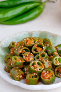 A dish of sliced korean chili peppers coated in a sweet ssamjang sauce and garnished with sesame seeds, served on a green ceramic plate. Whole green chilies are blurred in the background.