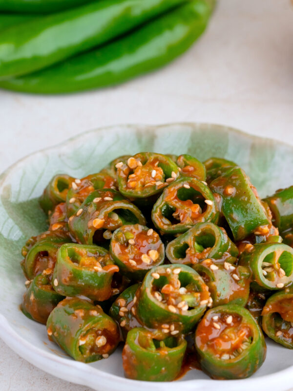 A dish of sliced korean chili peppers coated in a sweet ssamjang sauce and garnished with sesame seeds, served on a green ceramic plate. Whole green chilies are blurred in the background.