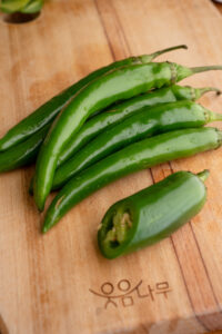 A group of fresh korean chili peppers rests on a wooden cutting board, alongside half a jalapeno pepper